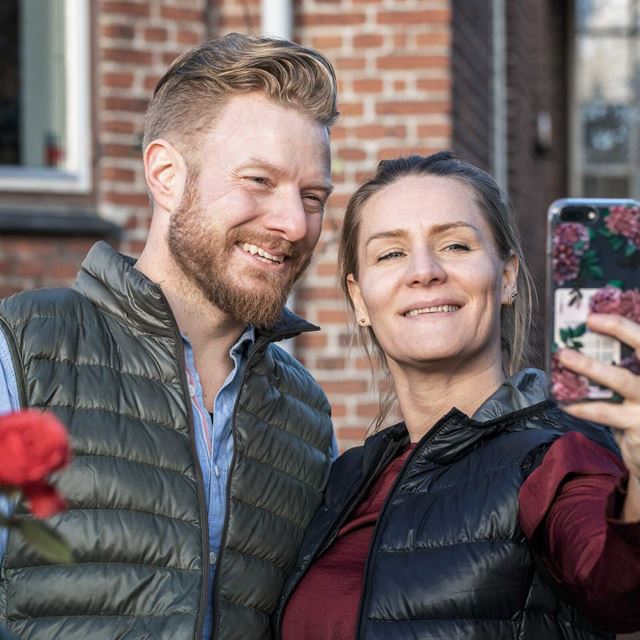 Et smilende par står udendørs foran et murstenshus og tager en selfie sammen.