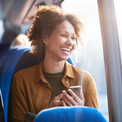 En kvinde sidder i en bus og smiler bredt, mens hun holder sin telefon og kigger ud af vinduet.