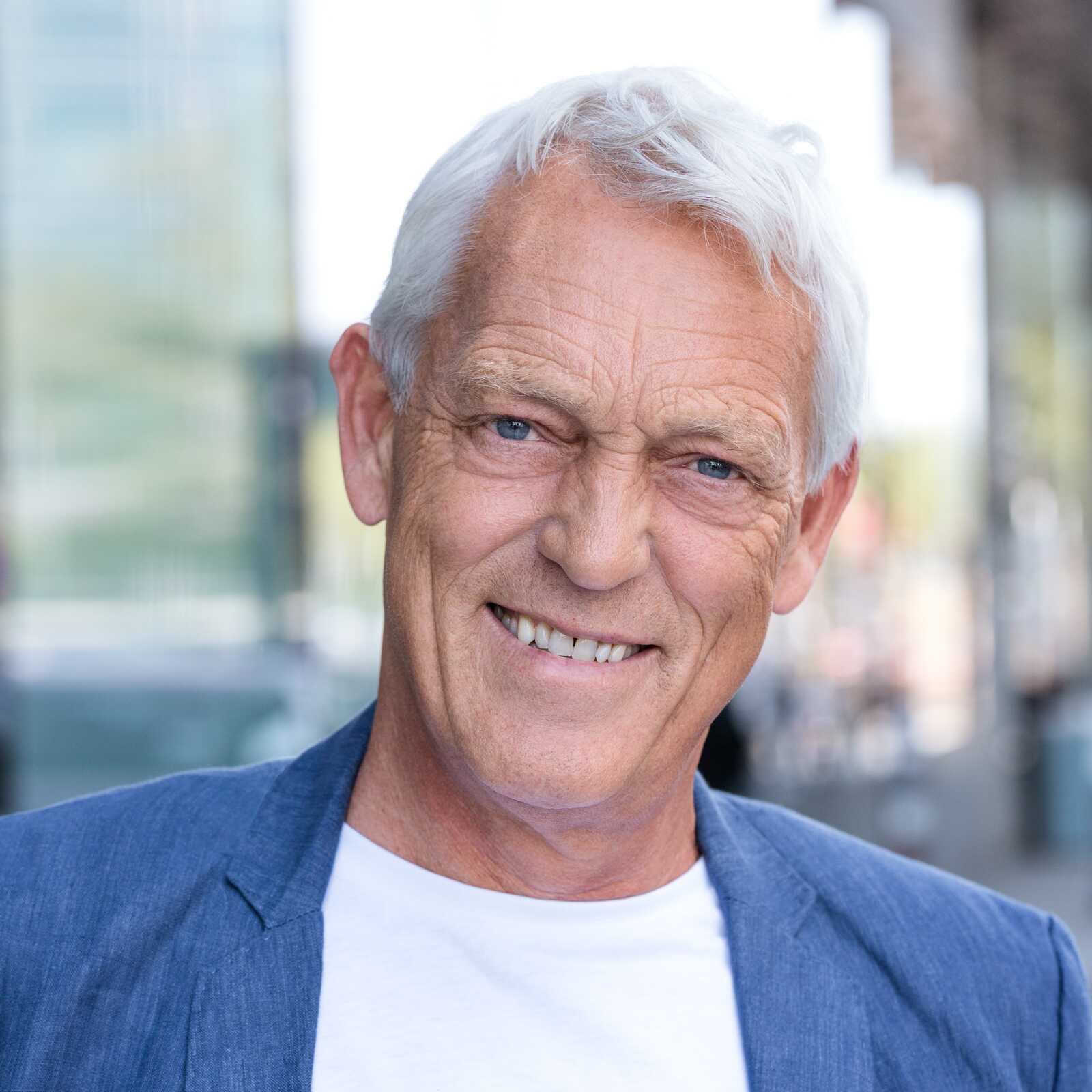 En ældre mand med hvidt hår smiler varmt til kameraet iført en blå blazer og en hvid t-shirt.