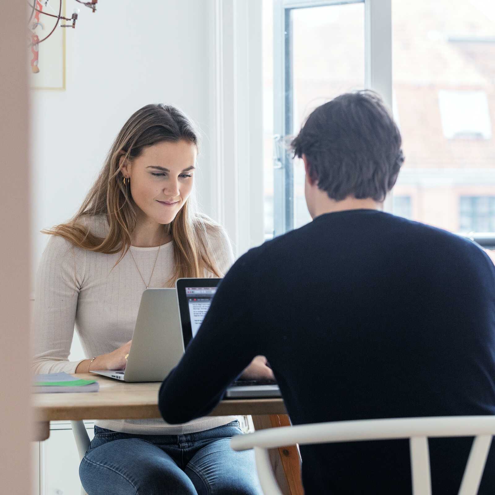 To studerende arbejder koncentreret ved et bord med deres bærbare computere, mens de sidder i et lyst rum.