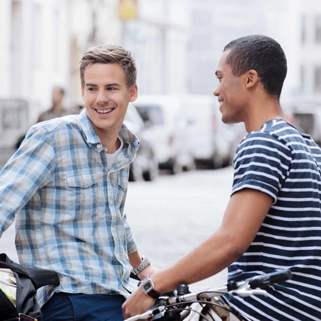 To unge mænd står og smiler, mens de snakker sammen på gaden. Den ene står ved sin cykel.
