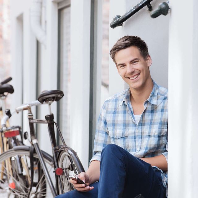 En ung mand i ternet skjorte sidder afslappet på en trappesten ved siden af cykler, smilende og kigger på sin telefon.
