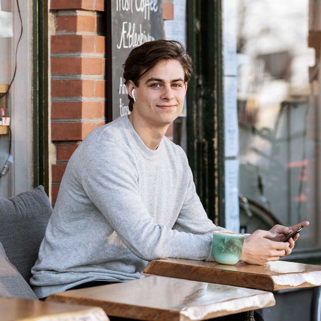 En ung mand sidder afslappet på en café med sin telefon og en kop kaffe ved siden af sig, nydende øjeblikket.