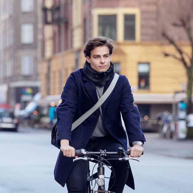 En ung mand cykler gennem byen, fokuseret og velklædt, med tasken hængende over skulderen.