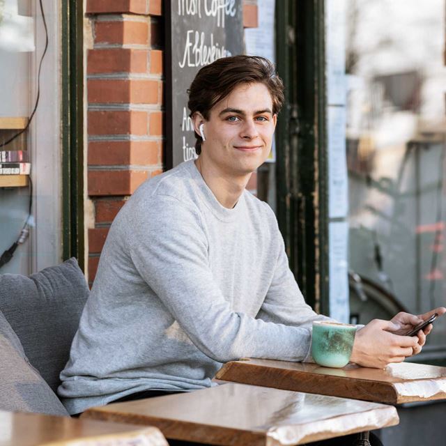 En ung mand sidder udenfor på en café og smiler til kameraet. Han har øretelefoner i og holder en telefon i hånden, mens der står en kop kaffe på bordet foran ham.