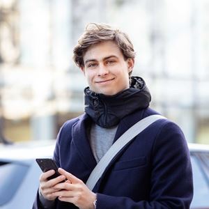 Ung mand iført mørk frakke smiler mod kameraet, mens han holder en telefon i hånden udendørs. Han har en afslappet og venlig fremtoning.