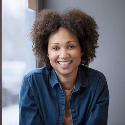 Ung kvinde med krøllet hår og denimskjorte smiler varmt og ser direkte på kameraet. Hendes udtryk udstråler venlighed og positivitet.