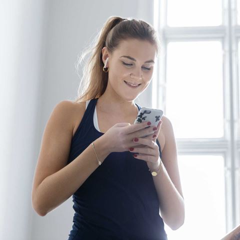En ung kvinde i træningstøj med hestehale smiler, mens hun kigger på sin telefon, og sollyset strømmer ind gennem vinduet bag hende.