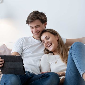 Ungt par sidder sammen i sofaen, griner og ser på en tablet, mens de er afslappede og smilende.