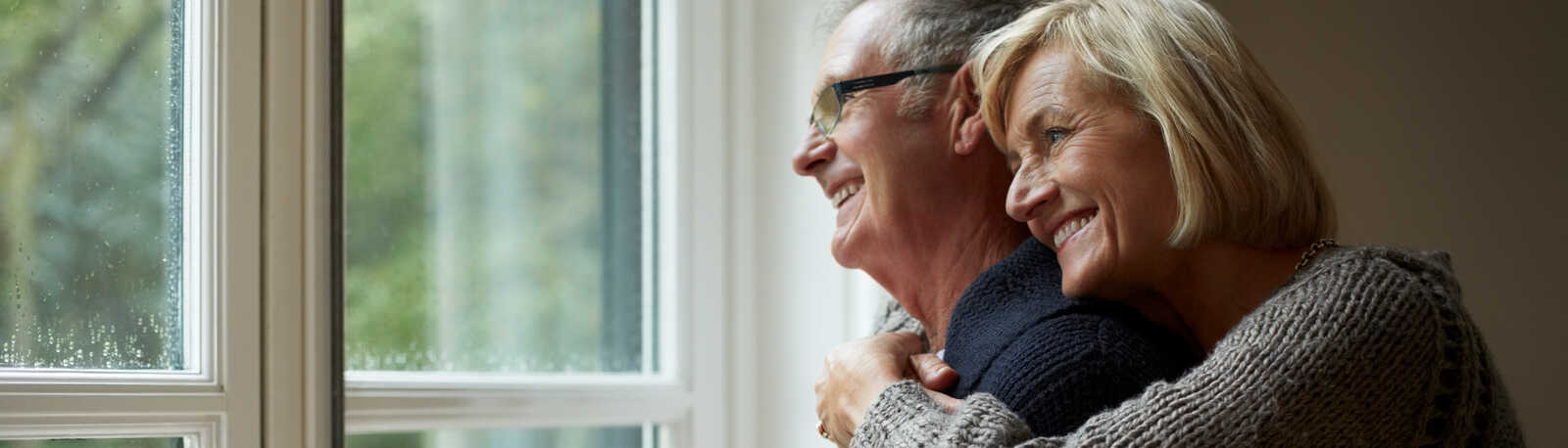 Ældre par står ved vinduet, kvinden krammer manden bagfra. Han holder en kop. Udendørs udsigt gennem vinduet, begge smiler varmt.