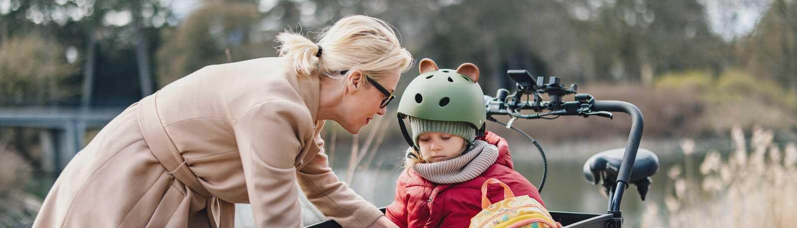 Mor hjælper sin datter med at spænde sikkerhedsselen i en ladcykel. Barnet bærer cykelhjelm og rygsæk, omgivelserne er udendørs med natur.