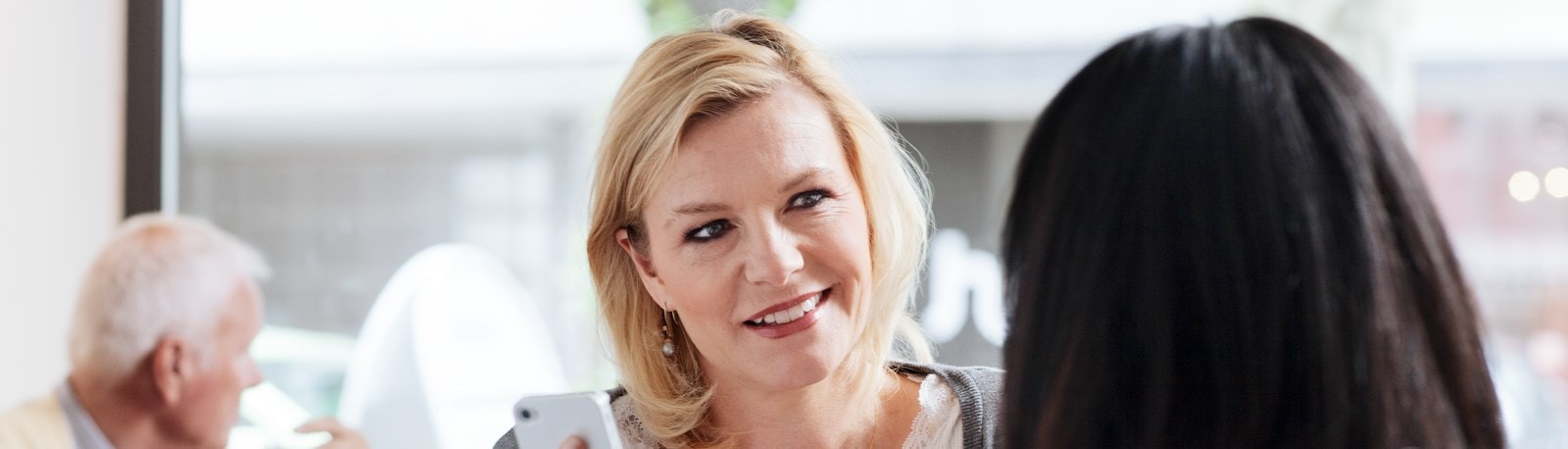 To kvinder hygger sig sammen på en café, begge smilende, mens de kigger på noget på deres mobiltelefon, der skaber en munter stemning.