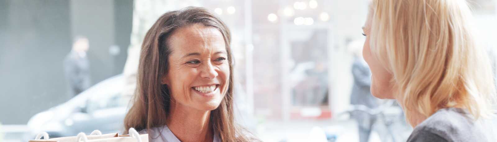 To kvinder smiler og snakker sammen på en café, omgivet af drikkevarer og shoppingposer.