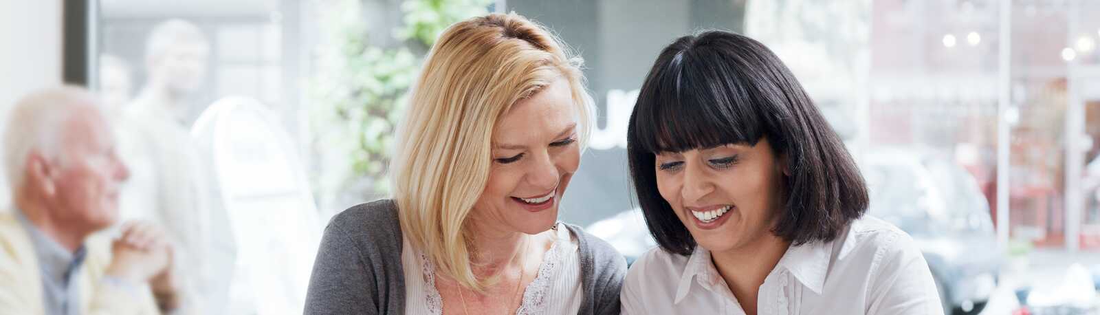 To kvinder sidder på en café og smiler, mens de kigger på noget på en telefon og nyder deres kaffe.