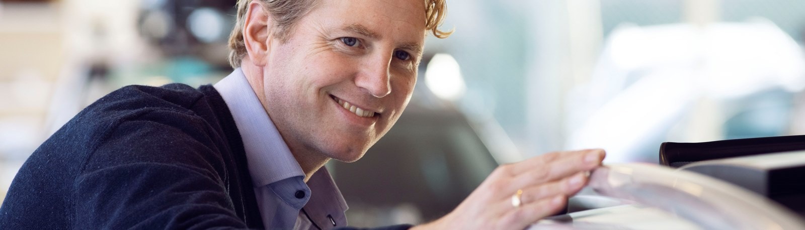 Smilende mand i forretningsdragt læner sig kærligt mod en ny bil i et showroom.