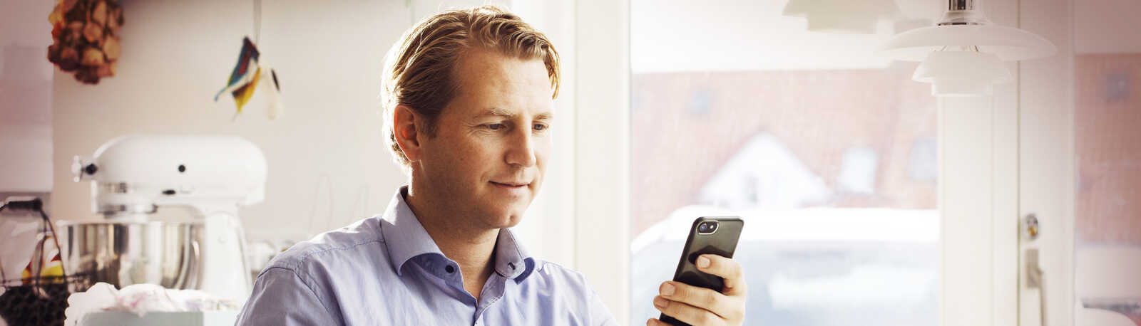 mand med en blå skjorte sidder i et køkken, med en kop kaffe og kigger på din telefon