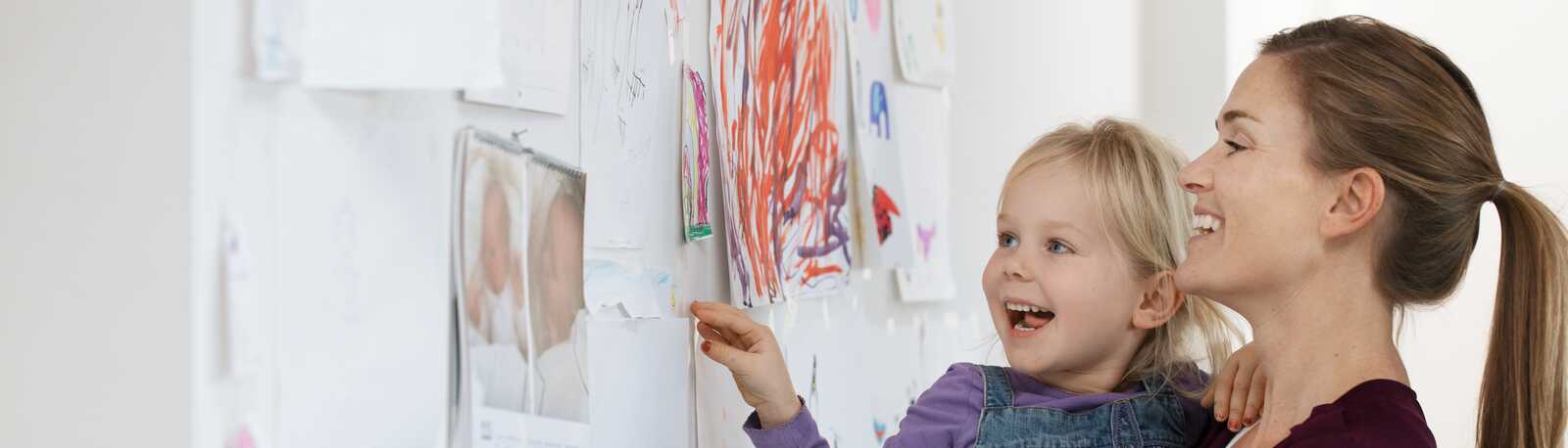 En mor og datter smiler, mens de ser på børnetegninger, der er hængt op på væggen.