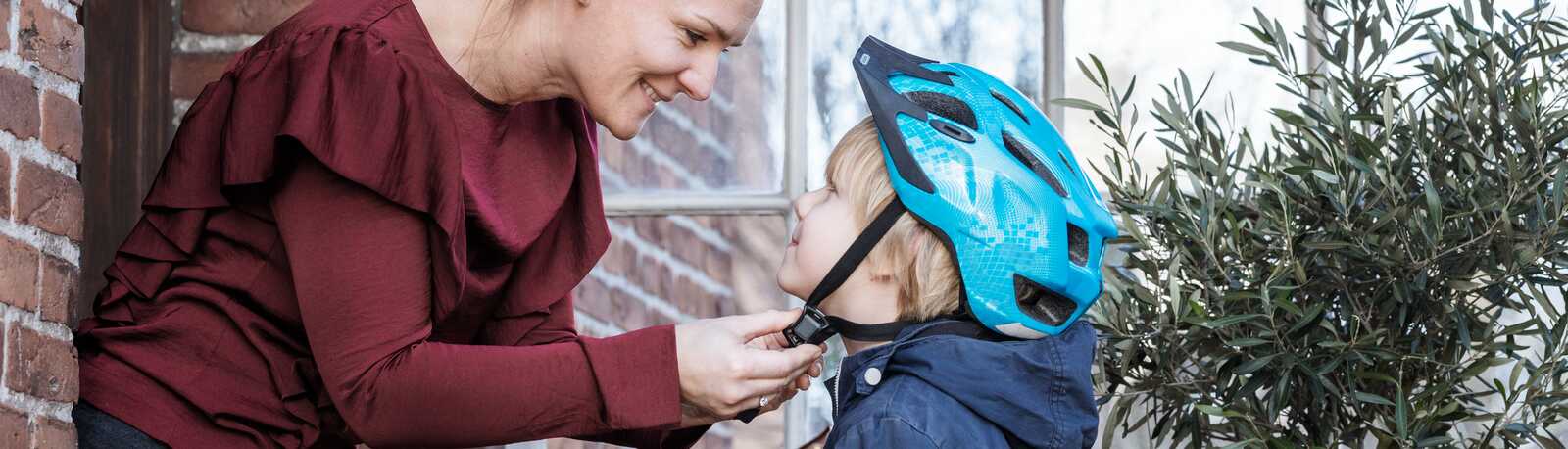 En mor fastgør sin søns cykelhjelm, mens de står udendørs foran et murstenshus og smiler til hinanden.