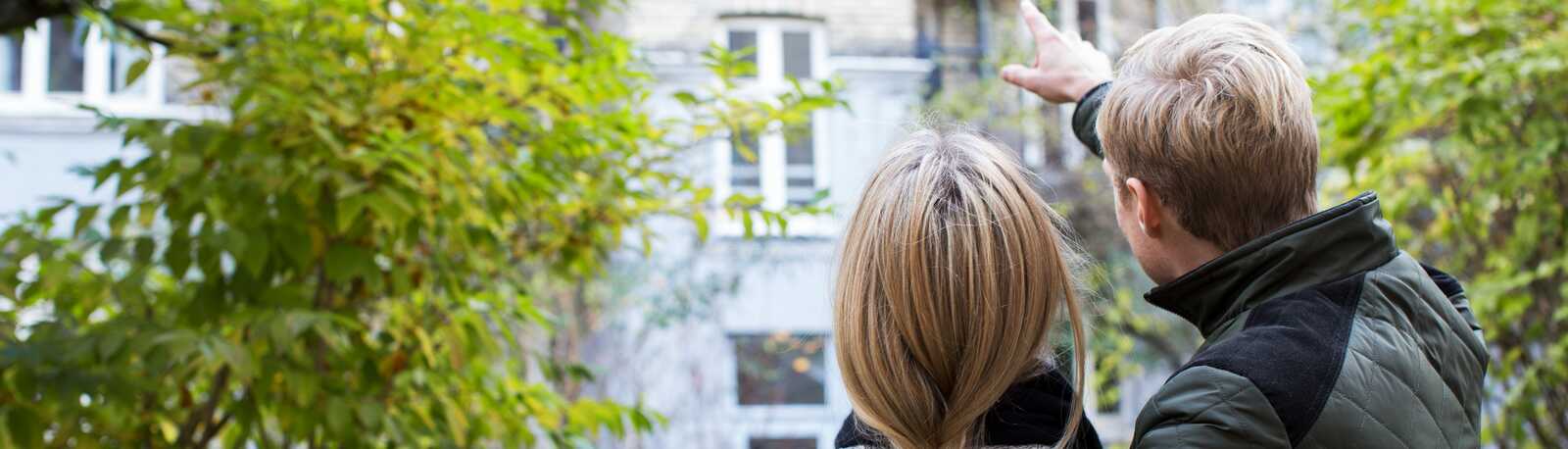 Et par står i en gårdhave og peger mod en lejlighedsbalkon, mens de kigger op på bygningen sammen.