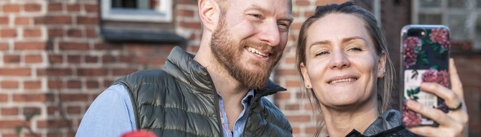Et smilende par står udendørs foran et murstenshus og tager en selfie sammen.