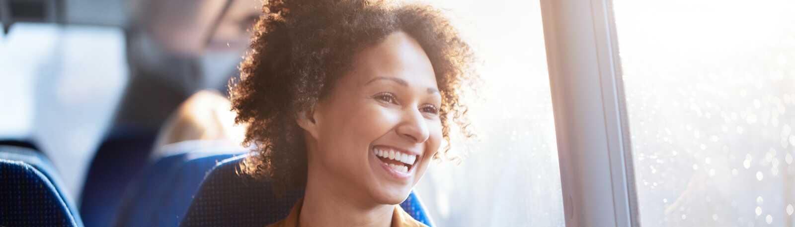 En kvinde sidder i en bus og smiler bredt, mens hun holder sin telefon og kigger ud af vinduet.