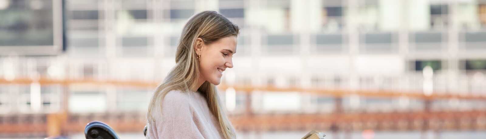 En ung kvinde sidder på en bænk ved vandet og smiler, mens hun læser en bog på en solrig dag.