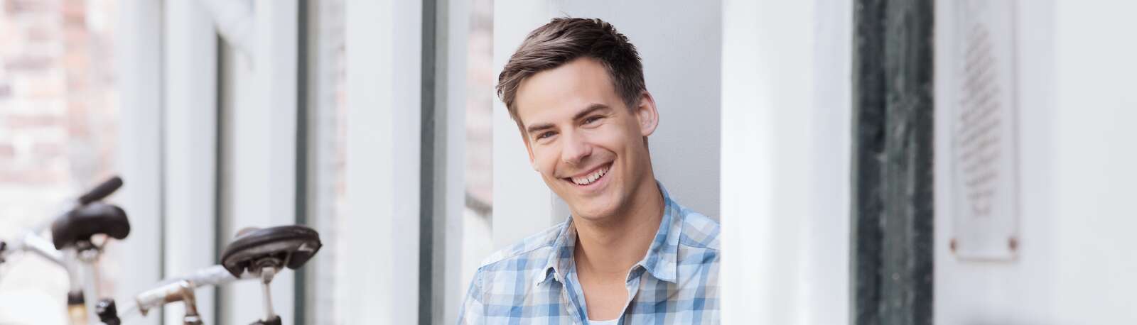 En ung mand i ternet skjorte smiler og kigger ind i kameraet, mens han sidder ved en cykel parkeret på gaden.