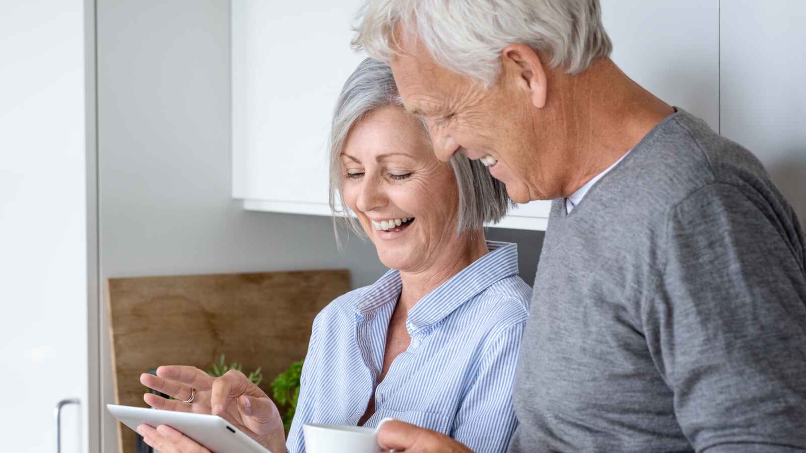 Parret smiler sammen, mens de kigger på en tablet i køkkenet. Kvinden holder en kop, og omgivelserne er lyse og moderne med en hyggelig atmosfære.