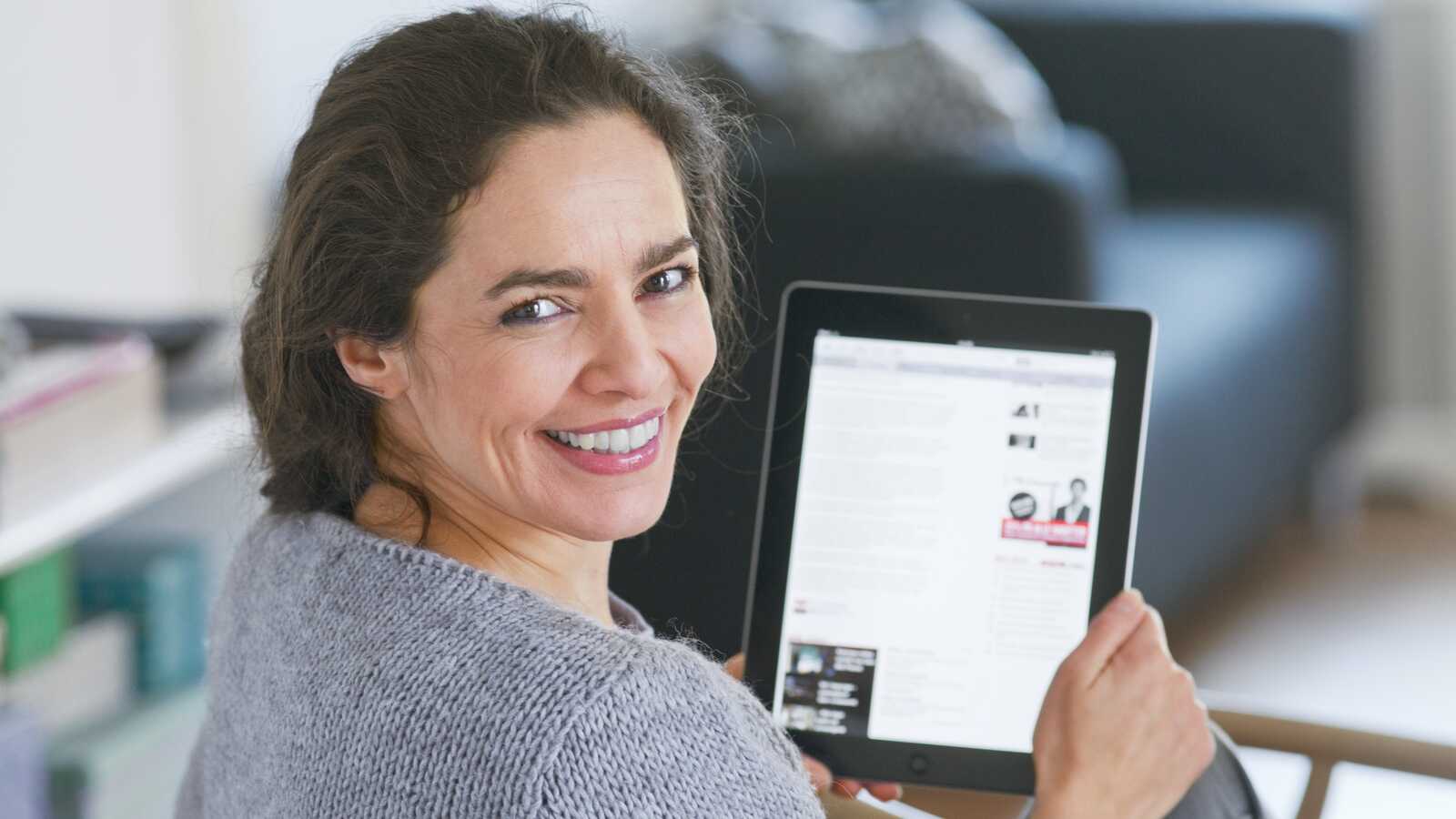 Kvinde sidder og smiler, mens hun holder en tablet, der viser en artikel. Indendørs omgivelser med en sofa og sløret baggrund.