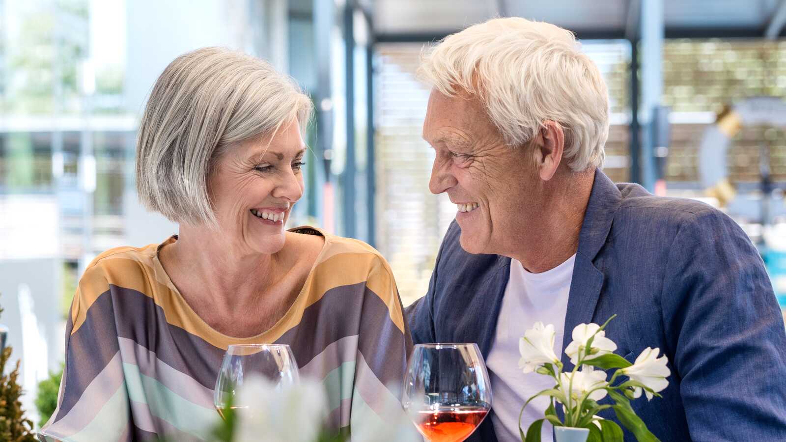 Ældre par sidder ved et restaurantbord, smiler til hinanden og nyder vin. Blomster på bordet og moderne omgivelser i baggrunden.
