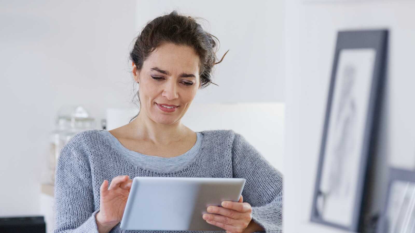 Kvinde i grå sweater står i køkkenet og kigger på en tablet. Baggrunden er lys med køkkenredskaber og glasbeholdere.