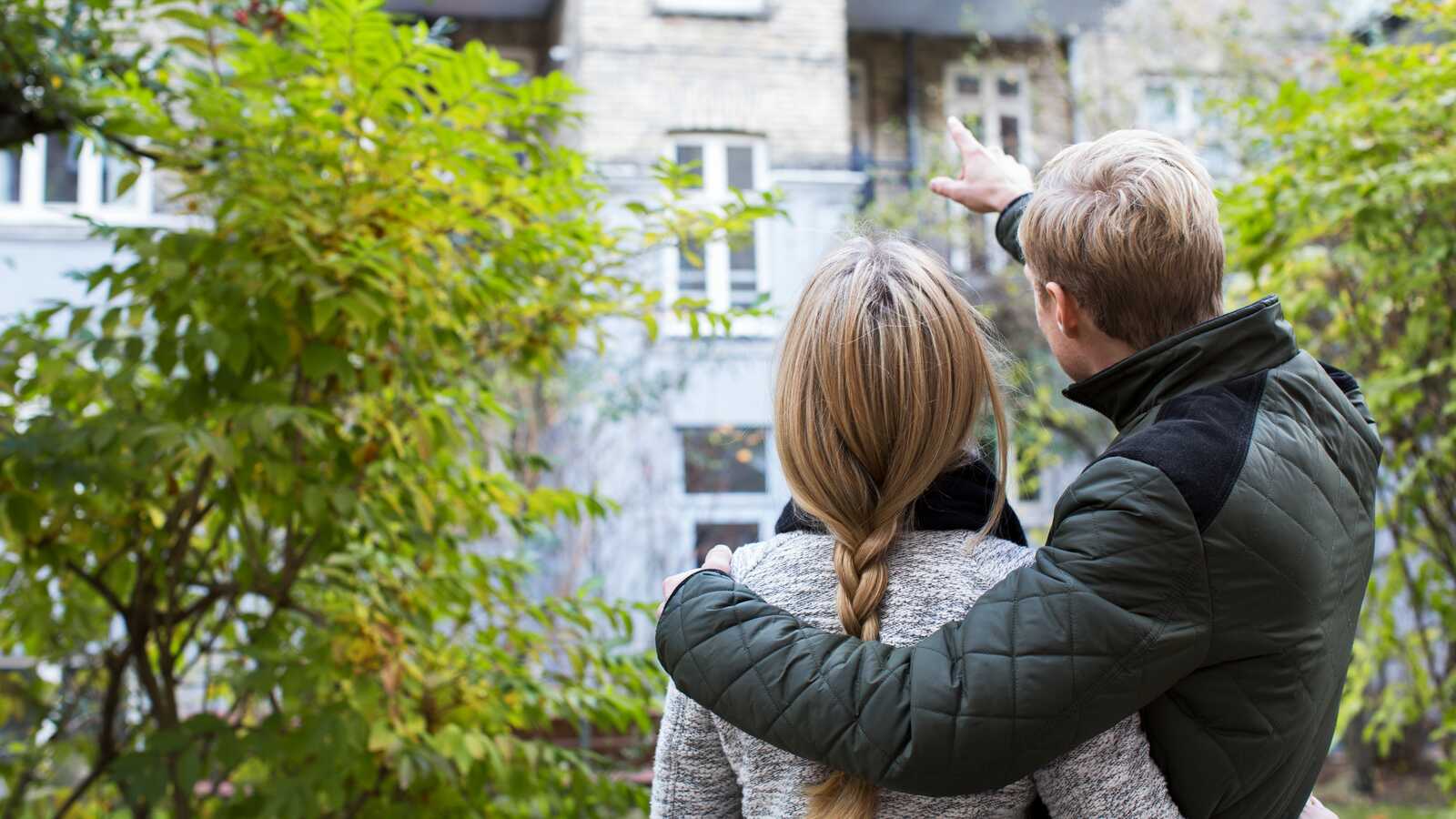 Et par står i en gårdhave og peger mod en lejlighedsbalkon, mens de kigger op på bygningen sammen.