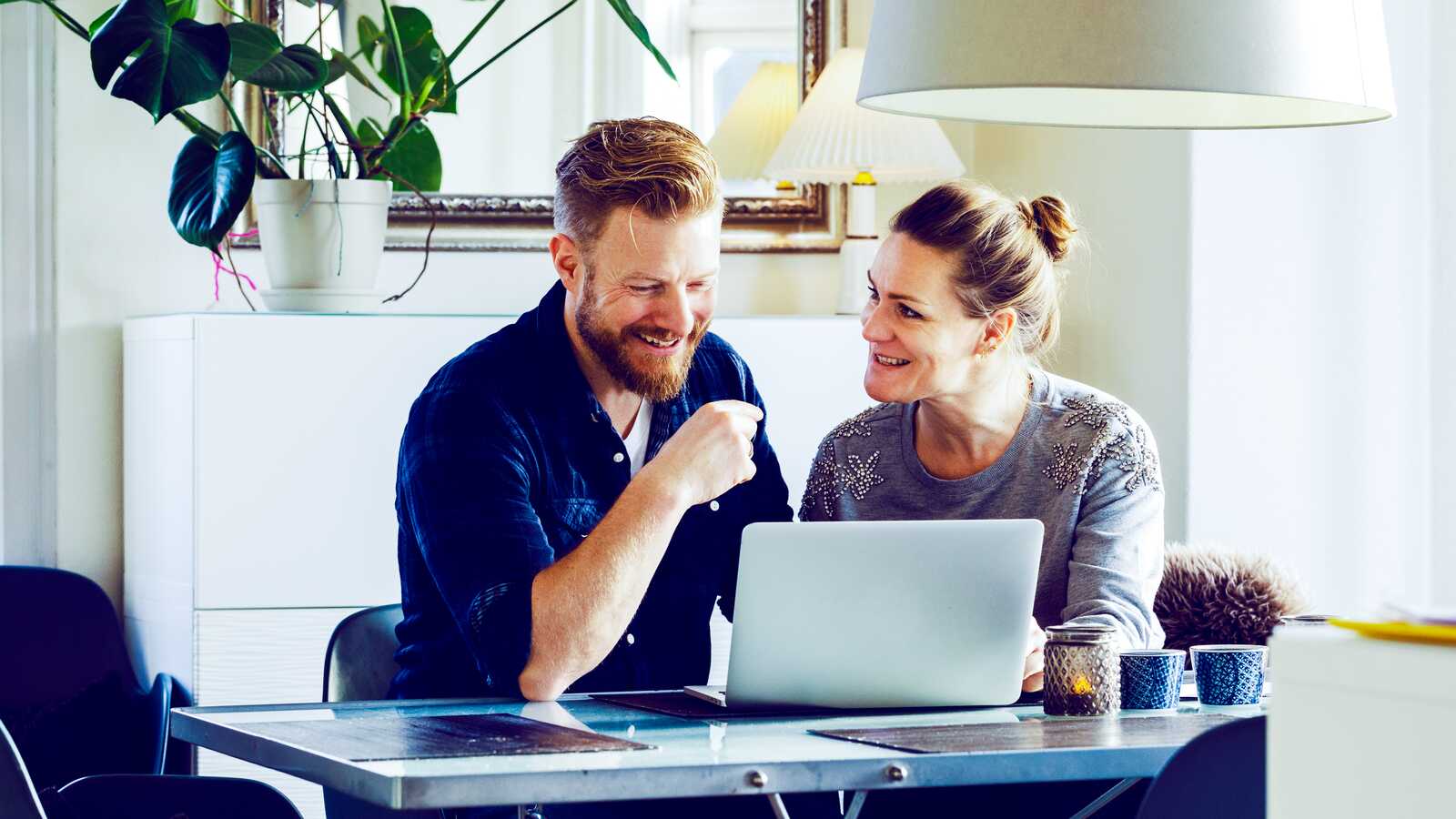 Et smilende par sidder ved et bord i stuen og arbejder sammen på en bærbar computer, mens de diskuterer noget.