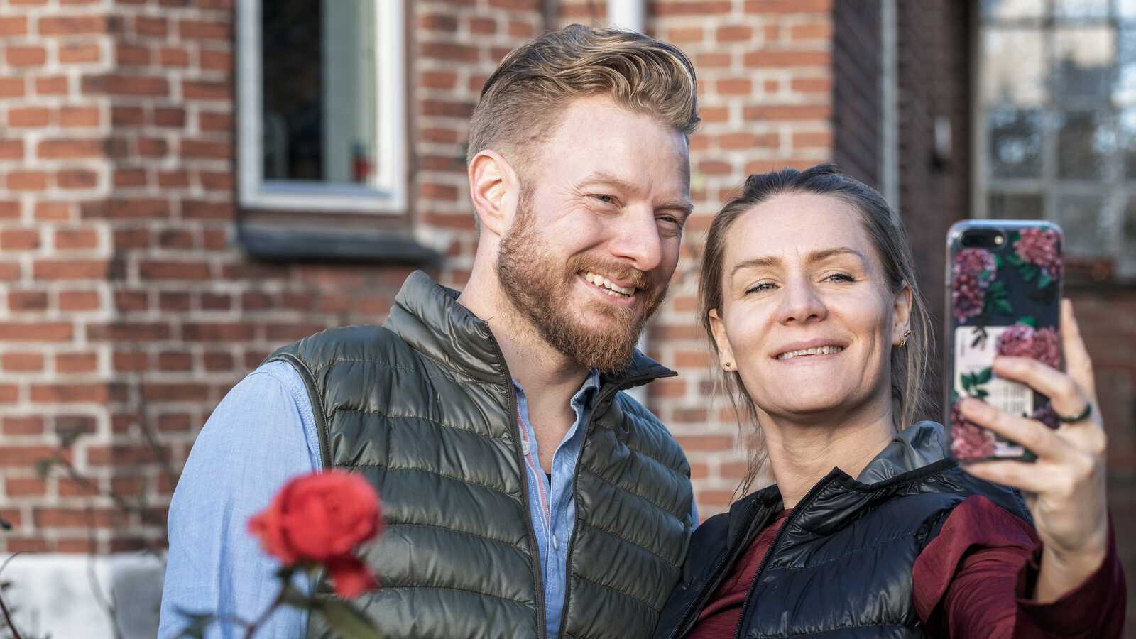 Et smilende par står udendørs foran et murstenshus og tager en selfie sammen.