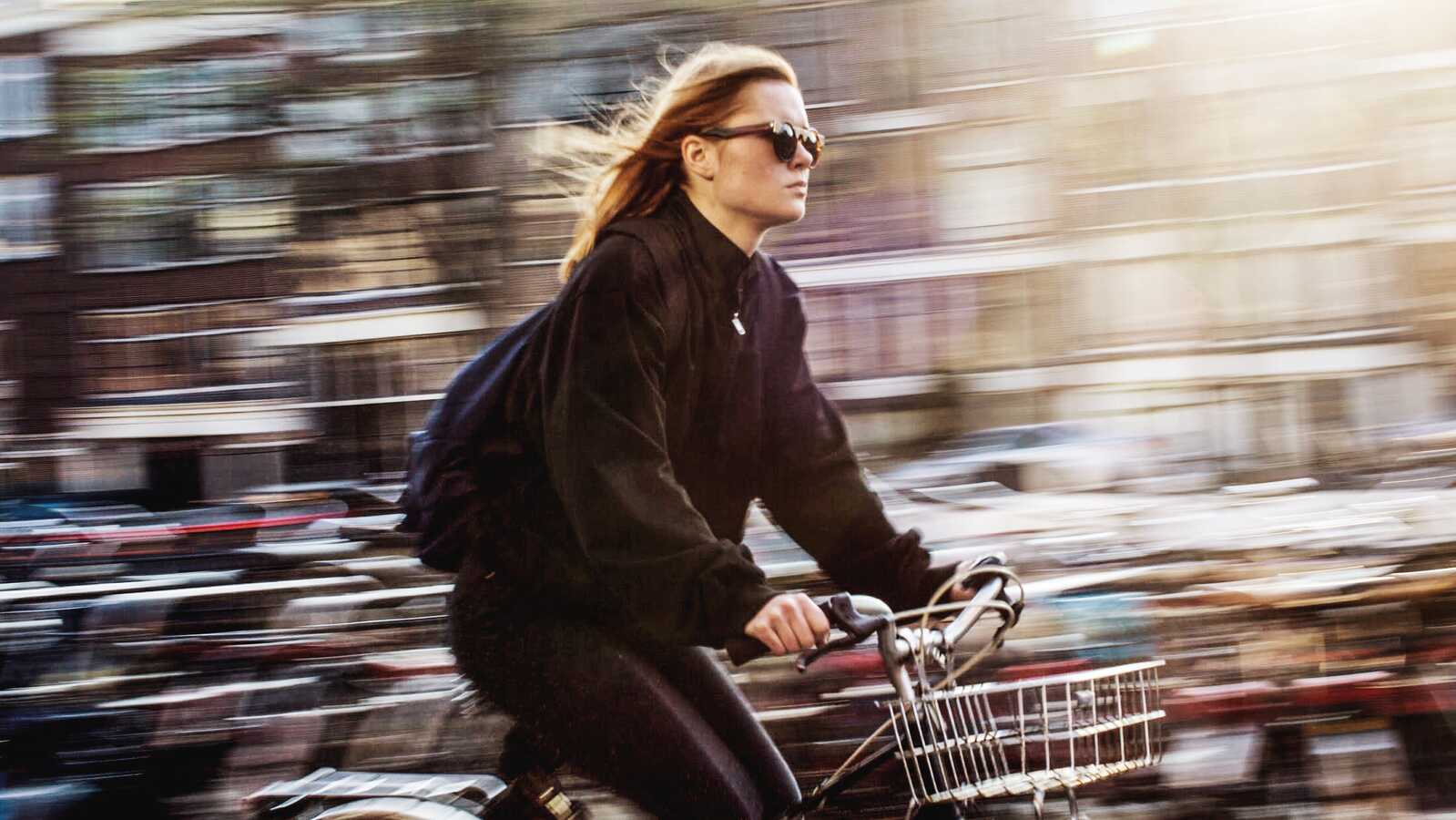 En kvinde iført solbriller cykler hurtigt gennem byen med vinden i håret på en solrig dag.