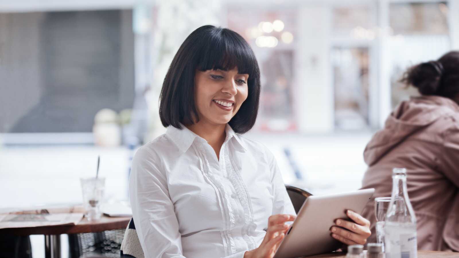 smilende kvinde sidder på cafe i en hvis skjorte med sin tablet og arbejder