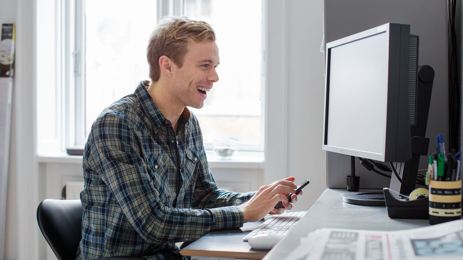 Begejstret ung mand smiler, mens han arbejder ved sin computer og holder sin telefon i hånden.