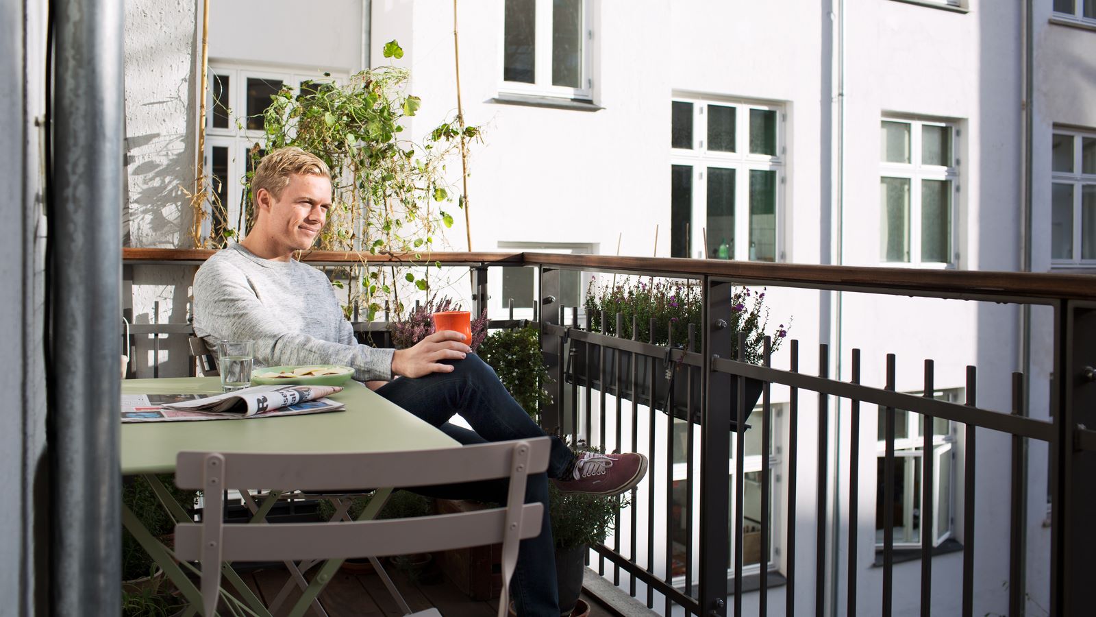 ung lyshåret mand sidder på sin altan med grønne havemøbler og drikker en kaffe