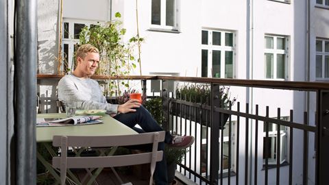 ung lyshåret mand sidder på sin altan med grønne havemøbler og drikker en kaffe