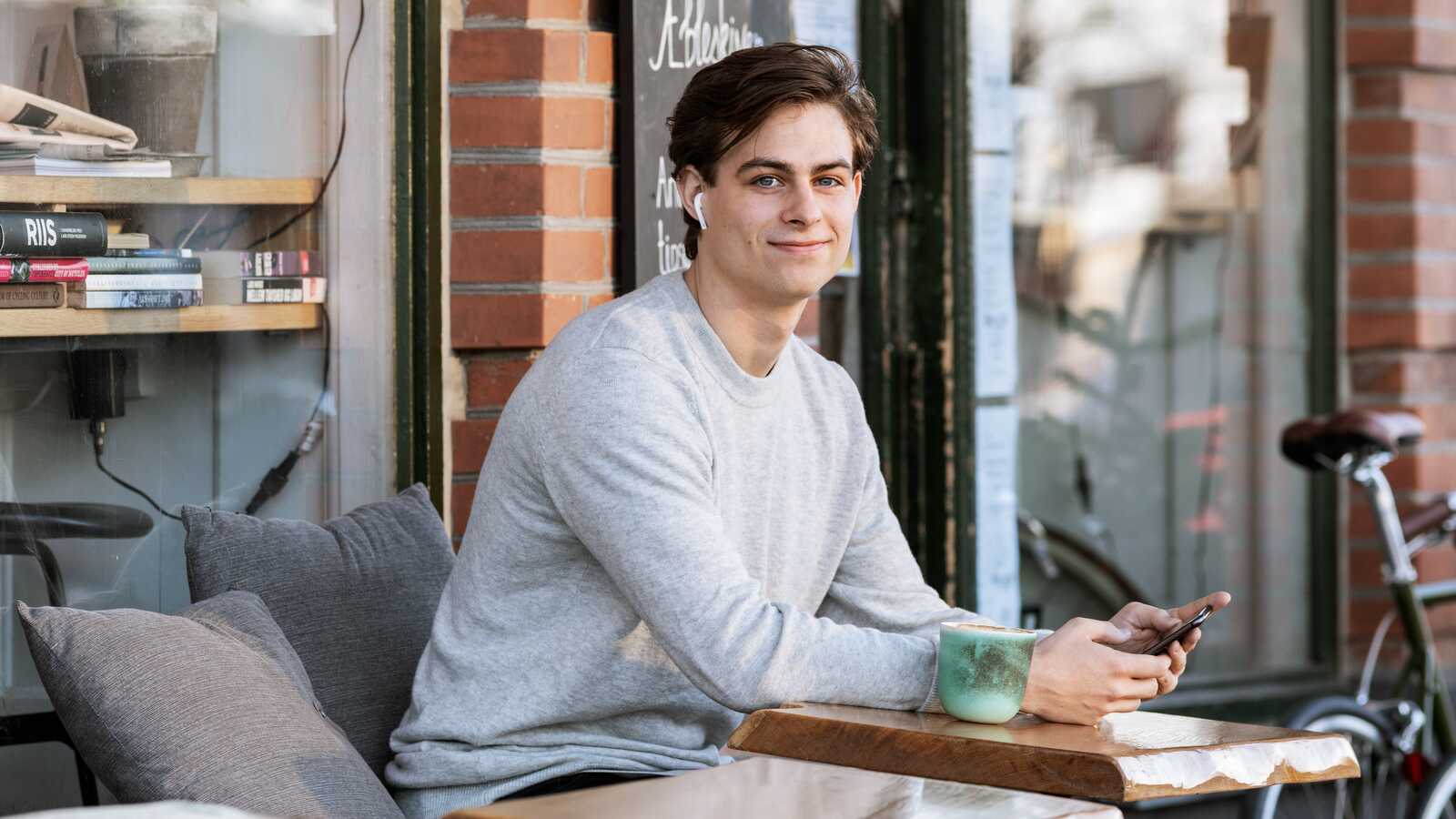 En ung mand sidder afslappet på en café med sin telefon og en kop kaffe ved siden af sig, nydende øjeblikket.